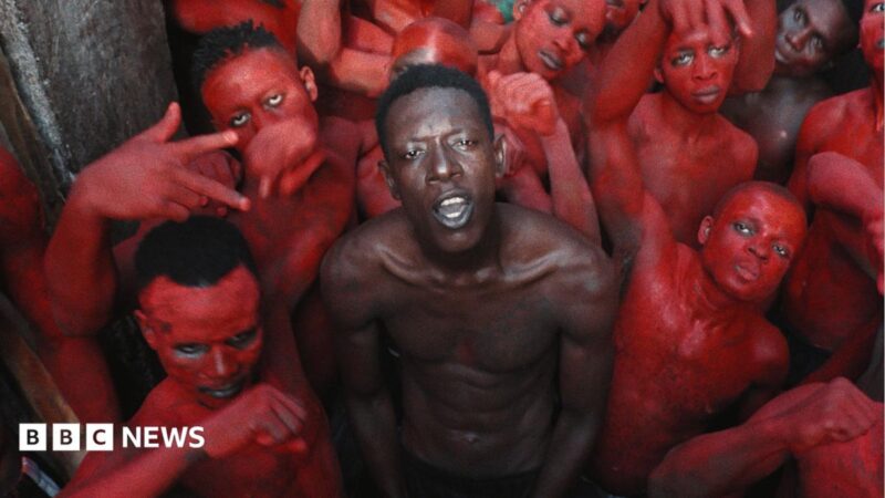 A topless Empereur stands in the centre of a group of young men, who are painted in red