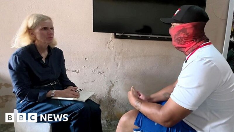 Orla Guerin speaks to a man wearing a black baseball cap and red scarf covering his face