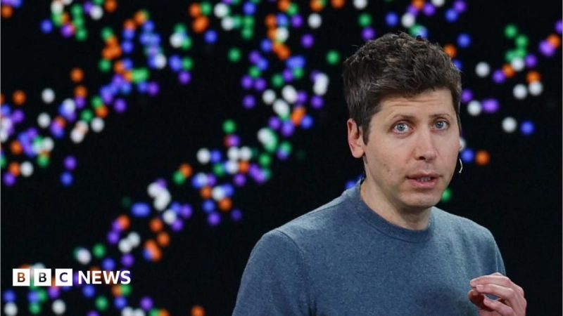 Sam Altman stands in front of a black background with many different-coloured dots. He's wearing a blue top and gesturing