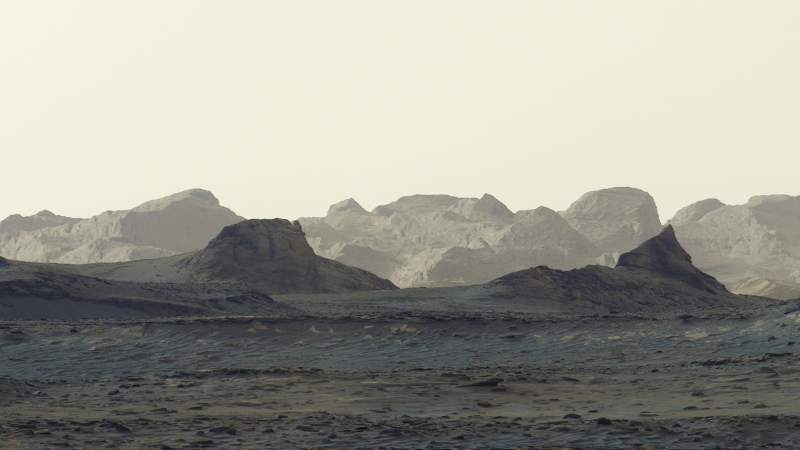 A stark, grayish-tan Martian landscape features jagged rock formations and rolling mountain ridges beneath a pale, hazy sky