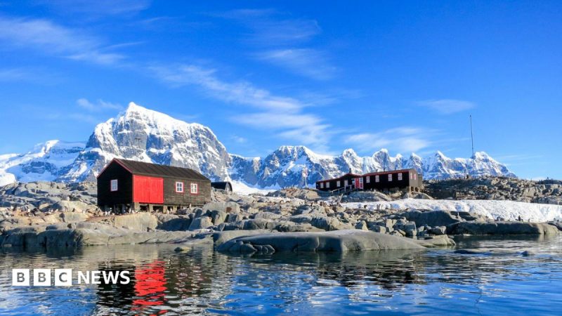 Two red and black huts sit on rocky ground beside calm Antarctic waters, with snow-covered peaks rising in the background
