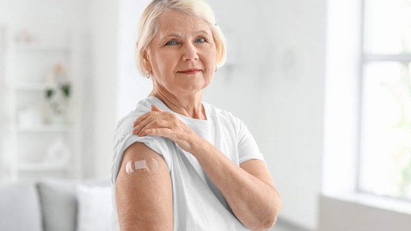 Older woman in a white shirt shows a small bandage on her upper arm after receiving a vaccination