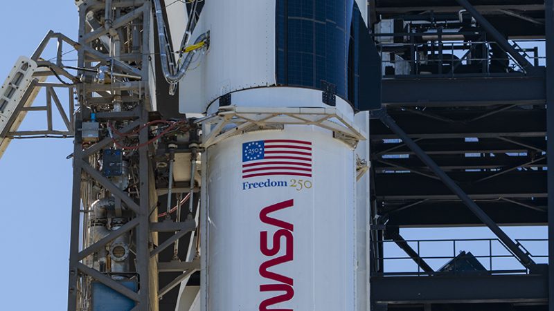 A close-up view of a SpaceX Crew Dragon spacecraft mounted atop a Falcon 9 rocket on the launch pad. The capsule and trunk are connected to ground support hardware, with NASA and mission insignia visible on the rocket’s exterior against a bright blue sky.