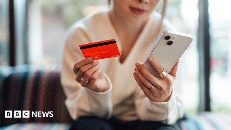 A woman sitting on a sofa and shopping online with her white phone while holding her red bank card in the other hand. She is