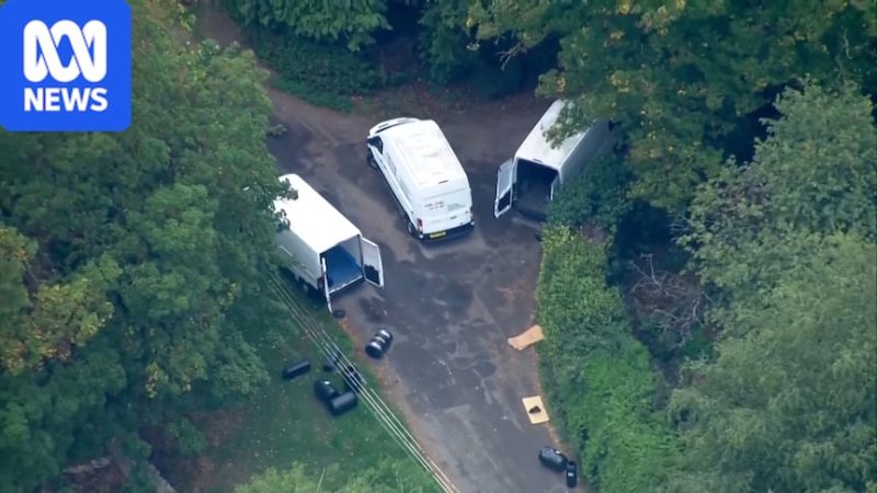 An aerial shot of three white vans, with two of their rear doors open, on a road near to multiple black barrels