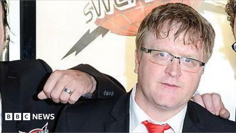 Mike Smith appearing at a film premiere is wearing a dark suit coat and red tie. The hands of two co-stars are seen on his