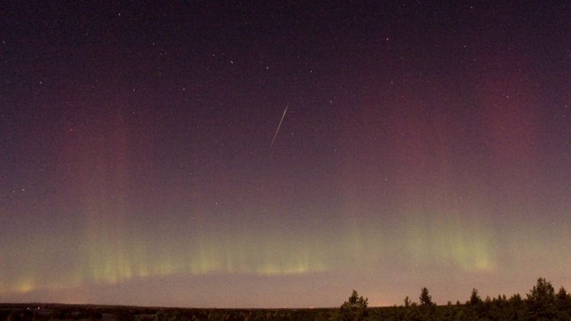 A lone shooting star streaks through a night sky riven with colorful aurora